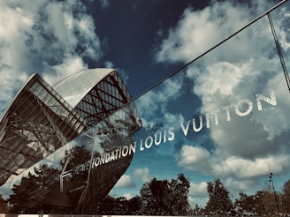 A modern glass building with a unique, curvilinear architectural design is partially reflected in a glass facade. The sky is cloudy with a mix of blue and white. The words 'Fondation Louis Vuitton' are prominently displayed on the glass.