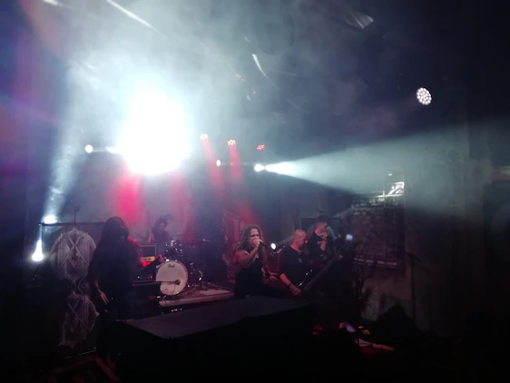 A band is performing on stage under dramatic lighting, which includes bright white and red spotlights. The lead singer is at the forefront, while other band members are playing musical instruments like a guitar and drums in the background. The stage is filled with smoke, adding to the intense and atmospheric ambiance.