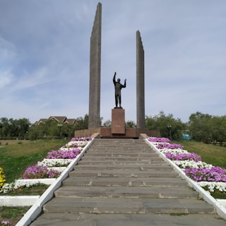 A monumental sculpture of a figure standing on a platform with outstretched arms, flanked by two tall vertical pillars. A stairway leads up to the statue, bordered by neatly arranged flowers in shades of pink, purple, and white. In the background, there is greenery and a few residential buildings.