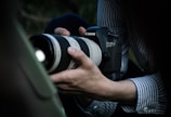 A person wearing a striped shirt is holding a Canon EOS 6D camera with a large zoom lens, capturing a subject. The setting appears to be outdoors with a slightly blurred background.