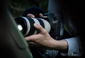 A person wearing a striped shirt is holding a Canon EOS 6D camera with a large zoom lens, capturing a subject. The setting appears to be outdoors with a slightly blurred background.