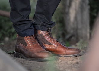 Brown leather boots with a lace-up design are worn with dark pants. The setting appears to be outdoors, with a blurred natural background that includes hints of greenery.