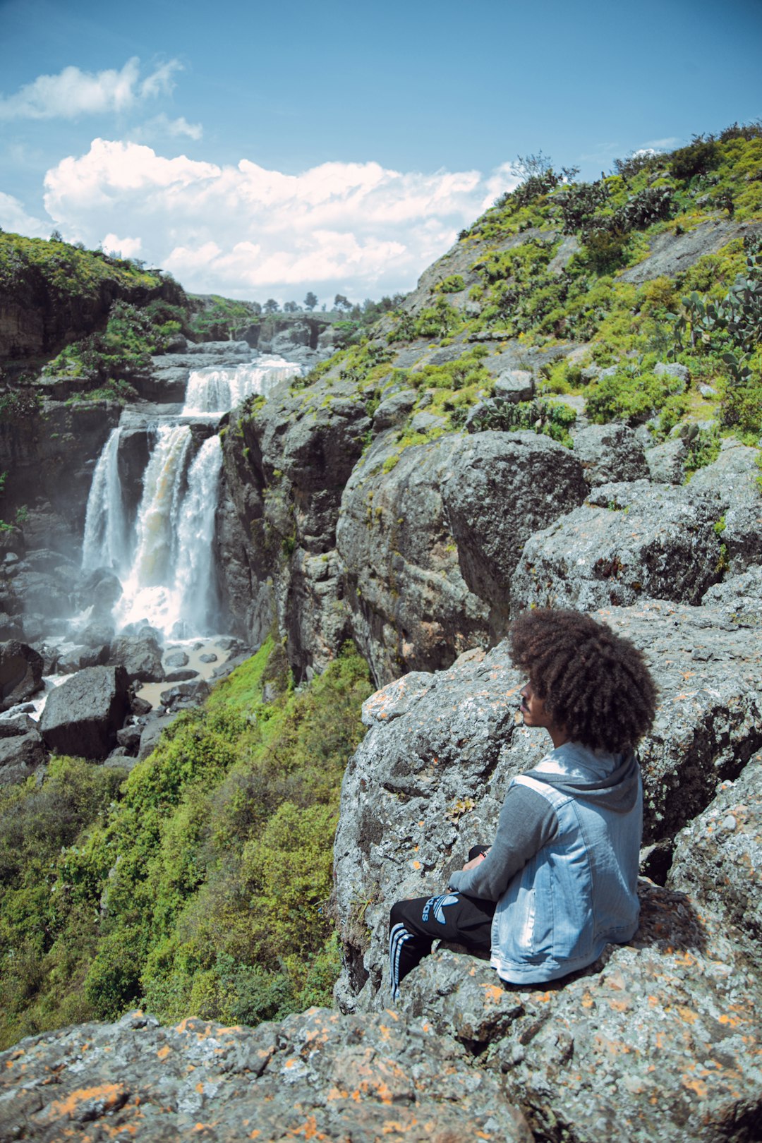 man in gray jacket sitting on rock looking at waterfalls during daytime