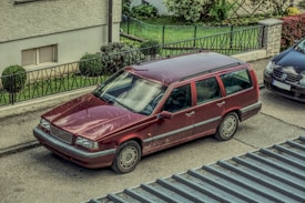 A red Volvo station wagon parked on a residential street next to a black Volkswagen car. The vehicles are situated beside a sidewalk with a metal railing and greenery visible in the background. The scene is viewed from above.