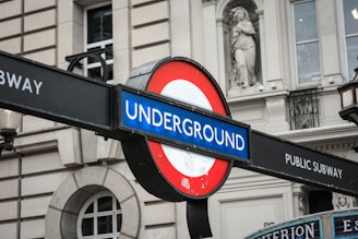 A classic London Underground sign showcasing a circular red and blue logo with the word 'UNDERGROUND' in white text. The sign is mounted on a metal pole, situated in front of a historical building facade with decorative stonework and windows.