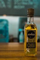 A bottle of chia oil labeled 'Aceite de Chia' with branding that includes the name 'Jevica.' The label also highlights that the oil is premium, edible, and 100% natural. The bottle is positioned on a wooden surface in front of an out-of-focus background featuring cool-toned colors.