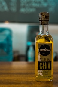 A bottle of chia oil labeled 'Aceite de Chia' with branding that includes the name 'Jevica.' The label also highlights that the oil is premium, edible, and 100% natural. The bottle is positioned on a wooden surface in front of an out-of-focus background featuring cool-toned colors.