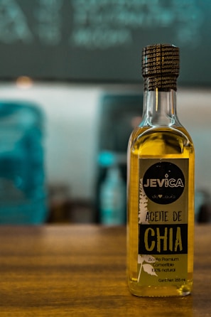 A bottle of chia oil labeled 'Aceite de Chia' with branding that includes the name 'Jevica.' The label also highlights that the oil is premium, edible, and 100% natural. The bottle is positioned on a wooden surface in front of an out-of-focus background featuring cool-toned colors.