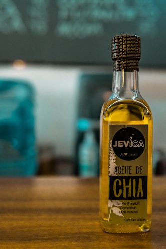 A bottle of chia oil labeled 'Aceite de Chia' with branding that includes the name 'Jevica.' The label also highlights that the oil is premium, edible, and 100% natural. The bottle is positioned on a wooden surface in front of an out-of-focus background featuring cool-toned colors.