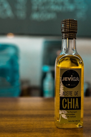 A bottle of chia oil labeled 'Aceite de Chia' with branding that includes the name 'Jevica.' The label also highlights that the oil is premium, edible, and 100% natural. The bottle is positioned on a wooden surface in front of an out-of-focus background featuring cool-toned colors.