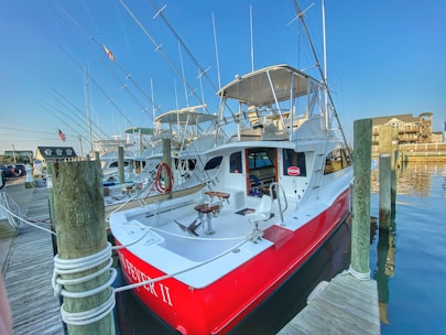 A red and white fishing boat docked at a marina, equipped with multiple fishing rods pointing upwards. The boat is tied to a wooden pier with ropes. Nearby, other boats are visible, and residential buildings are in the background. An American flag is also present.