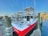 A red and white fishing boat docked at a marina, equipped with multiple fishing rods pointing upwards. The boat is tied to a wooden pier with ropes. Nearby, other boats are visible, and residential buildings are in the background. An American flag is also present.