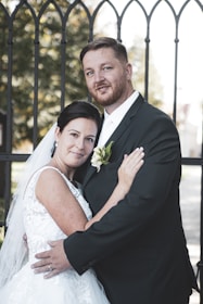 A couple, dressed formally with the woman in a white wedding dress and the man in a black suit, pose affectionately in front of an ornamental wrought iron gate. The woman has a veil and holds her partner closely, while the man gently embraces her with one hand on her back. There is lush greenery visible through the gate, hinting at a park or garden setting, and soft lighting enhances the intimate atmosphere.