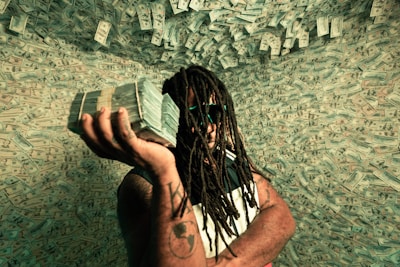 A person with long dreadlocks and sunglasses is holding a large stack of cash against a background filled with dollar bills. The setting creates a surreal atmosphere with money covering the walls.