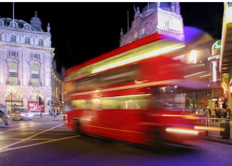 A red double-decker bus moves rapidly across a busy city street at night, creating a motion blur effect. Surrounding buildings are illuminated with bright lights, showcasing classic architecture. Neon signs and advertisements add vibrancy to the scene, while pedestrians are visible on the sidewalk.