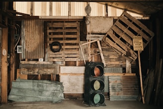 A rustic interior filled with various wooden pallets, corrugated metal sheets, and a hanging metal hook. A vintage traffic light stands upright at the center, adjacent to a warning sign about a buried petroleum pipeline. The walls and floor are made of rough, weathered wood, contributing to an industrial and somewhat cluttered atmosphere.