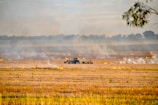 A field with scattered fires emitting smoke, and a tractor working among the flames. The background shows a horizon lined with trees under a partly cloudy sky. The scene indicates an agricultural practice involving controlled burning.