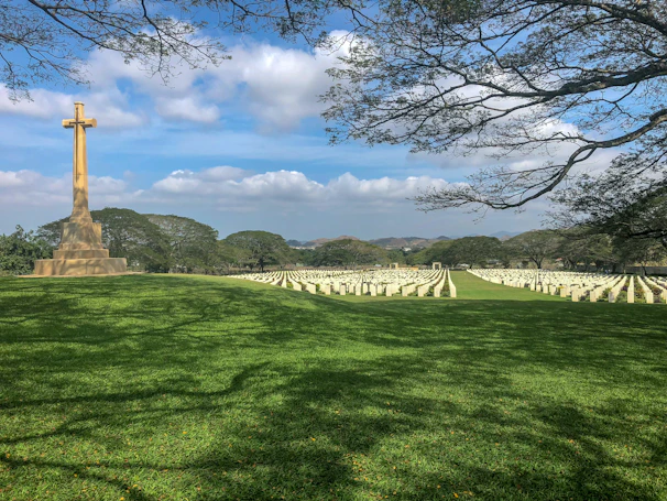 A large cemetery with rows of white headstones neatly arranged on a vast green lawn. A prominent stone cross stands to the left, with distant hills and a blue sky in the background. Leafless branches frame the image from the top.