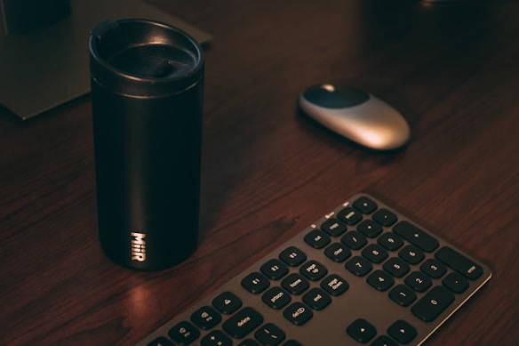 A black travel mug is on a wooden desk, accompanied by a keyboard and a computer mouse. The lighting is dim, creating a cozy atmosphere.