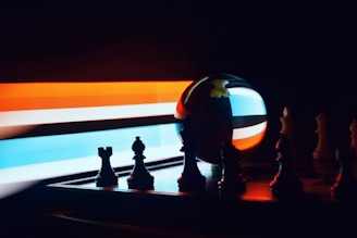 A chessboard with scattered chess pieces, primarily silhouetted against a blurred background of streaking colored lights. A reflective sphere mirrors the colors and the shapes of the pieces, adding a dynamic visual effect.
