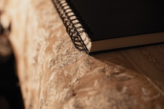 A close-up of a spiral-bound notebook resting on a surface with ornate, textured fabric. The lighting casts a warm, soft glow on both the notebook and the fabric, creating gentle shadows.