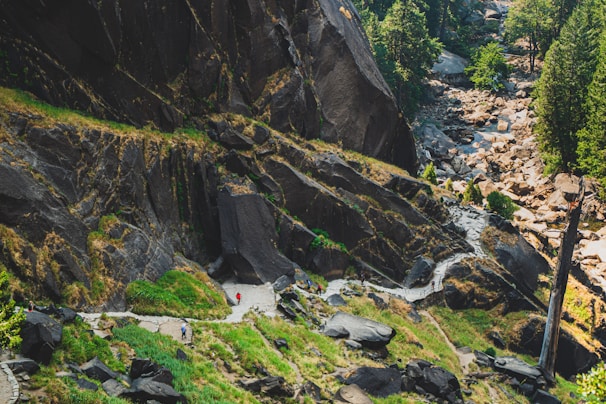 A winding path is carved into the lush landscape, bordered by steep rocky cliffs and tall, green trees. The scene includes a small group of people hiking along the trail, providing a sense of scale to the massive rocks surrounding them. The pathway leads through a vibrant green area, with patches of grass and shrubs scattered throughout the rough terrain.