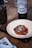 A plate with sliced meat displayed on a red sauce and garnished with small cucumbers. A bottle of organic Torrontés wine is placed nearby alongside a jar of pickles. The setting includes a folded napkin with cutlery on a wooden table.