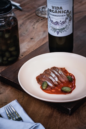 A plate with sliced meat displayed on a red sauce and garnished with small cucumbers. A bottle of organic Torrontés wine is placed nearby alongside a jar of pickles. The setting includes a folded napkin with cutlery on a wooden table.