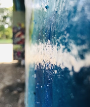 A close-up view of a concrete surface with a textured layer of blue paint, partially peeling. The background reveals an out-of-focus, colorful graffiti on another wall, and there are hints of a bright, sunlit area with greenery.
