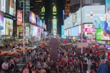 A bustling city scene filled with a vibrant crowd of people, surrounded by skyscrapers adorned with bright, colorful billboards and advertisements. Traffic flows with numerous yellow taxis and cars in the streets. The environment is lively with diverse individuals engaging and capturing the essence of the urban atmosphere.