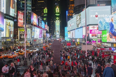 A bustling city scene filled with a vibrant crowd of people, surrounded by skyscrapers adorned with bright, colorful billboards and advertisements. Traffic flows with numerous yellow taxis and cars in the streets. The environment is lively with diverse individuals engaging and capturing the essence of the urban atmosphere.