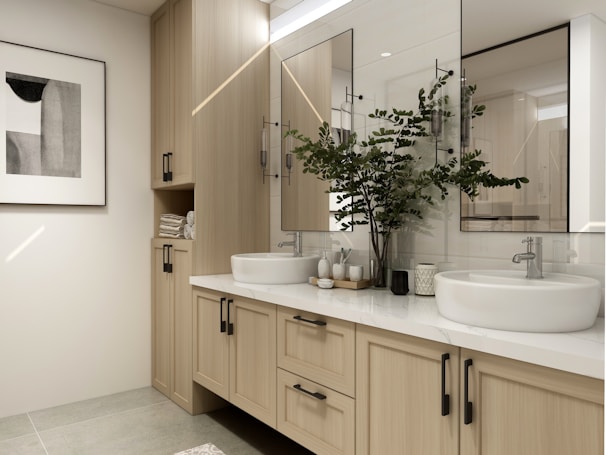 A modern bathroom vanity setup features double sinks with sleek chrome faucets, placed on a light wood cabinet with black handles. Above the sinks are large mirrors, and to the side, a potted plant adds a touch of nature. On the left, a minimalistic piece of black and white abstract art is mounted on the wall. Towels are neatly stored in an open section of the cabinetry, complementing the clean and organized aesthetic.