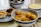 A collection of traditional Asian dishes is arranged on a table, including a bowl of yellow noodles with fish balls, a plate containing two egg tarts, and two bowls of desserts or soups. The dishes are served in blue and white ceramic bowls and plates.