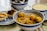A collection of traditional Asian dishes is arranged on a table, including a bowl of yellow noodles with fish balls, a plate containing two egg tarts, and two bowls of desserts or soups. The dishes are served in blue and white ceramic bowls and plates.