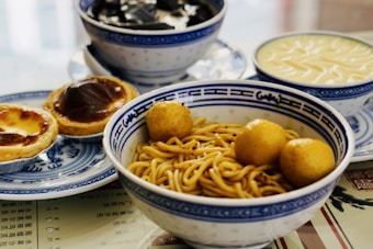 A collection of traditional Asian dishes is arranged on a table, including a bowl of yellow noodles with fish balls, a plate containing two egg tarts, and two bowls of desserts or soups. The dishes are served in blue and white ceramic bowls and plates.