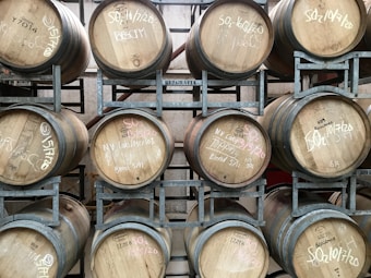 Several wooden barrels are arranged in a metallic rack. Each barrel has text written on it in white and pink chalk, indicating dates or specific identifiers. The barrels are stacked in rows, and the environment suggests a storage or cellar setting for aging beverages such as wine.