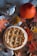 A freshly baked pie with a decorative braided lattice crust sits on a wooden surface surrounded by autumn leaves, a small pumpkin, acorns, and a metal coffee pot on a fabric cloth.