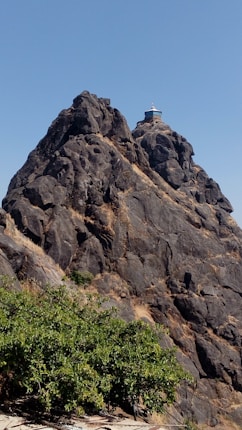 A rugged, rocky mountain peak with a small structure, possibly a temple or shrine, perched at the top. The mountain is steep and composed of dark grey rocks with some patches of dry grass. In the foreground, there is a green leafy bush or small tree growing from the rocky base.