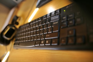 A close-up view of a black computer keyboard on a wooden desk with soft lighting. The perspective is slightly angled, focusing on the keys and showing a blurred background. There is also a computer mouse partially visible on the left side of the image.