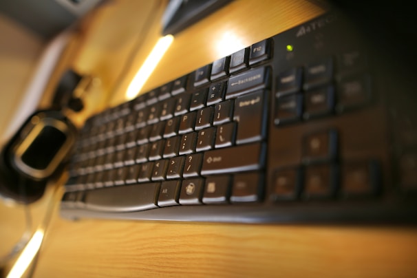 A close-up view of a black computer keyboard on a wooden desk with soft lighting. The perspective is slightly angled, focusing on the keys and showing a blurred background. There is also a computer mouse partially visible on the left side of the image.