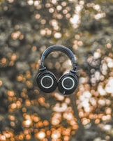 A pair of black over-ear headphones is suspended in mid-air against a backdrop of blurred lights. The background has a bokeh effect with warm tones, creating an abstract and artistic atmosphere.