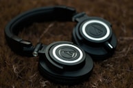 A pair of black over-ear headphones rests on a textured brown surface. The headphones feature a circular logo on each earcup, surrounded by silver and black detailing. The lighting is soft, creating a subdued atmosphere.