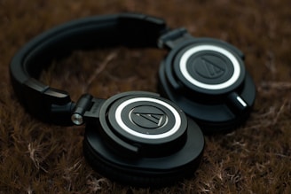 A pair of black over-ear headphones rests on a textured brown surface. The headphones feature a circular logo on each earcup, surrounded by silver and black detailing. The lighting is soft, creating a subdued atmosphere.