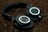 A pair of black over-ear headphones rests on a textured brown surface. The headphones feature a circular logo on each earcup, surrounded by silver and black detailing. The lighting is soft, creating a subdued atmosphere.