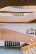 The image features modern architectural design with multiple levels of wooden slatted panels forming a staircase. The interior is spacious with large windows allowing natural light to illuminate the area. The wooden textures create a warm atmosphere, and the open layout presents a clean, minimalist style.