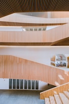 The image features modern architectural design with multiple levels of wooden slatted panels forming a staircase. The interior is spacious with large windows allowing natural light to illuminate the area. The wooden textures create a warm atmosphere, and the open layout presents a clean, minimalist style.