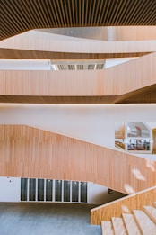 The image features modern architectural design with multiple levels of wooden slatted panels forming a staircase. The interior is spacious with large windows allowing natural light to illuminate the area. The wooden textures create a warm atmosphere, and the open layout presents a clean, minimalist style.
