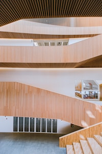 The image features modern architectural design with multiple levels of wooden slatted panels forming a staircase. The interior is spacious with large windows allowing natural light to illuminate the area. The wooden textures create a warm atmosphere, and the open layout presents a clean, minimalist style.