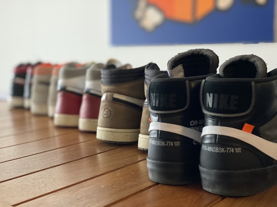A row of various Nike sneakers extends diagonally across a wooden floor, each pair slightly different in color and design. The image is focused on the back view, showcasing the brand and detailing on the heel tabs prominently. The background is blurred, containing a blue and orange abstract shape.
