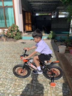 A young boy is riding a small bicycle with training wheels in a tiled courtyard. The bicycle is adorned with black and orange colors, and the boy is wearing a light purple shirt, black shorts, and sneakers. In the background, there is a house with wooden doors and large windows, surrounded by plants and flowers.